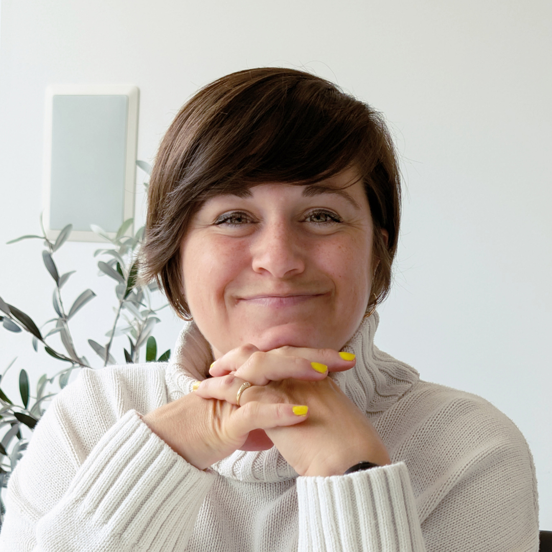 Une personne aux cheveux bruns courts et au vernis à ongles jaune, Valérie Noël, est assise à une table, le menton posé sur ses mains jointes, et sourit à l'appareil photo. Une plante feuillue se trouve à l'arrière-plan.