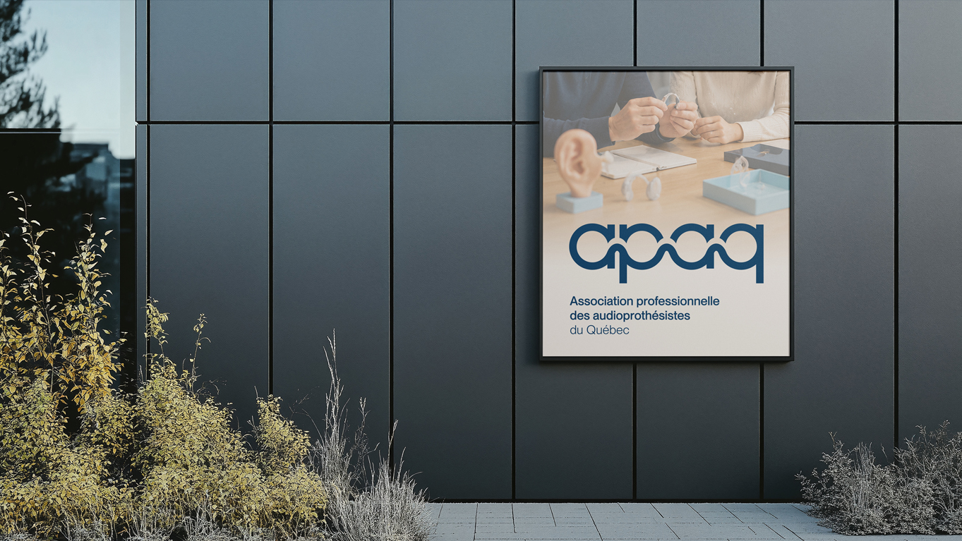 Affiche de l'Association professionnelle des audioprothésistes du Québec sur le mur sombre d'un immeuble, représentant des appareils auditifs, des maquettes d'oreilles et des mains manipulant ces appareils.